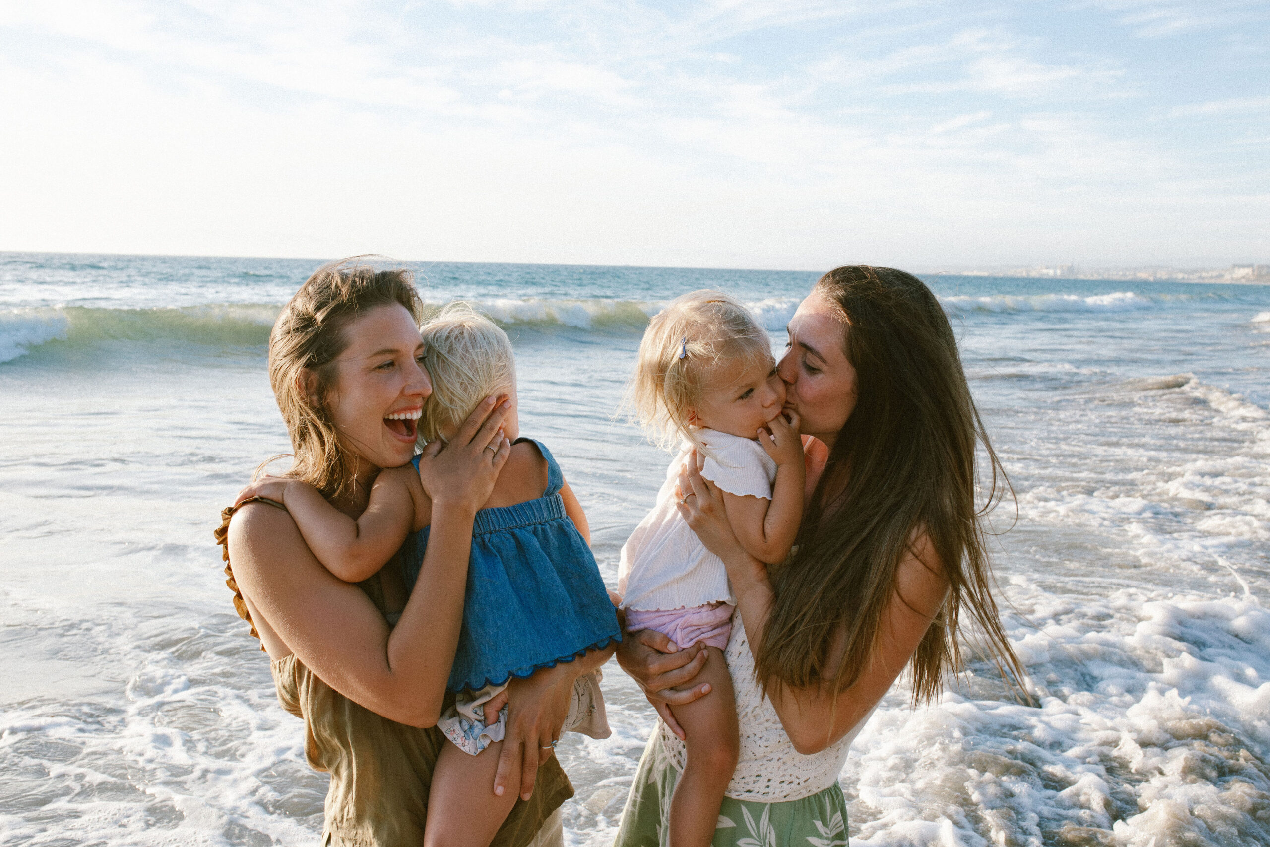 Two mothers holding their toddler daughters at the water's edge, one laughing, one kissing her child