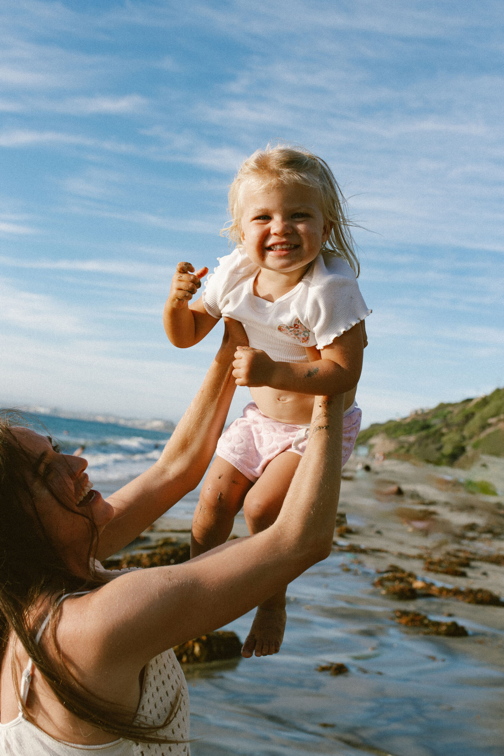 A mother lifting her laughing toddler daughter against a blue sky at the beach