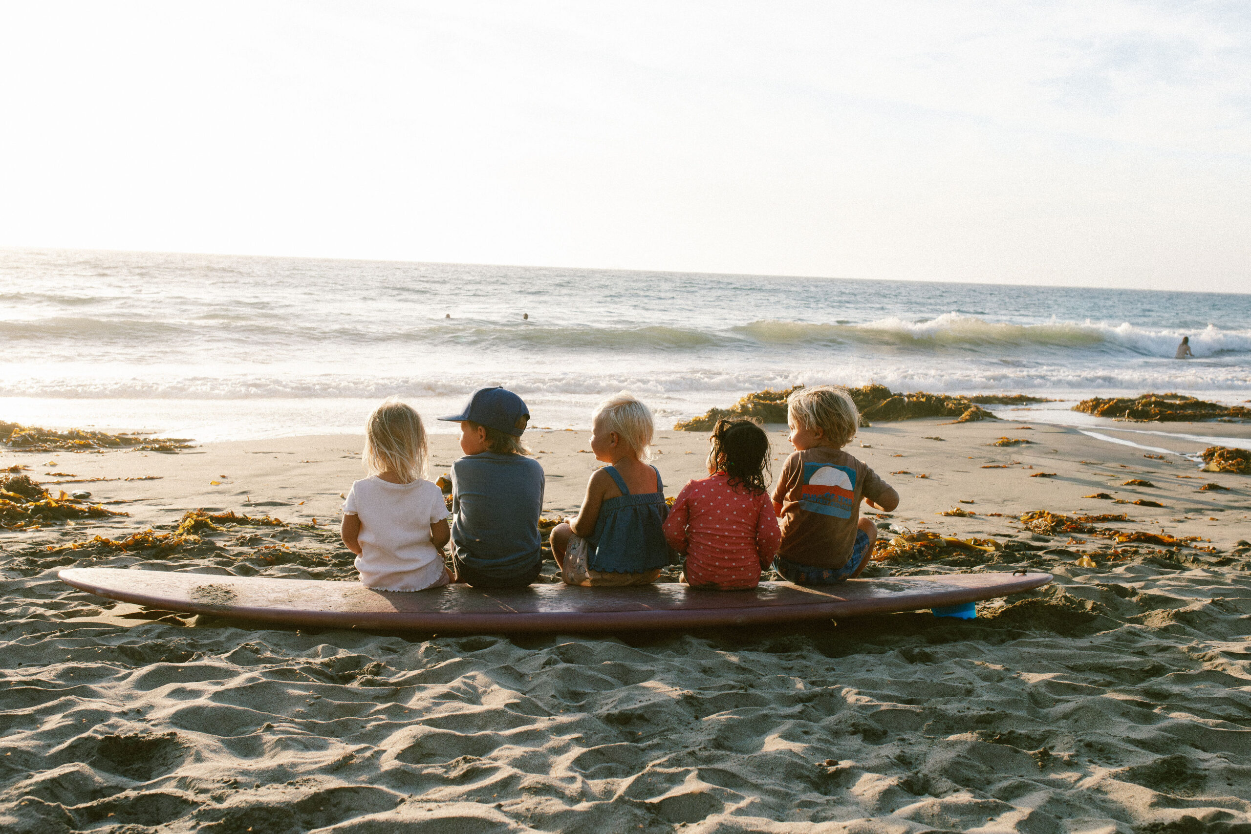 Five young children sitting in a row on a longboard, watching the surf at golden hour